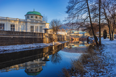 Historisches Kurgebäude mit grüner Kuppel spiegelt sich im Fluss vor verschneiten Ufern bei Bad Kissingen – erholsames Ziel für Silvester Busreisen zum Jahreswechsel