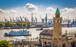 Hafenpanorama mit Dampfschiff, Menschen am Ufer und Kranen im Hintergrund.