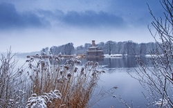 Winterlandschaft mit See, schneebedeckten Bäumen und einem Gebäude im Hintergrund.