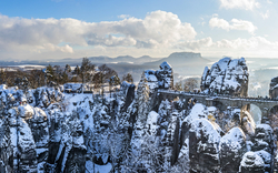 Verschneite Felsformation mit Brücke und bewaldeter Landschaft im Hintergrund.