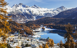 Bergdorf und See in Alpenlandschaft im Herbst mit schneebedeckten Gipfeln im Hintergrund.