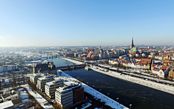 Ansicht einer verschneiten Stadt mit Fluss und Kirchtürmen im Hintergrund.