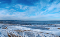 Verschneite Küstenlandschaft mit Leuchtturm am Horizont unter blauem Himmel.