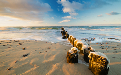 Wellen brechen an einem sandigen Strand mit Holzpfählen bei Sonnenuntergang.