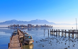 Hölzerner Steg mit Booten auf einem See, Berge im Hintergrund, viele Wasservögel.