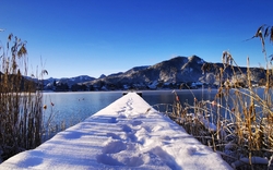 Verschneiter Steg am See vor Bergen und Schilf im Winter.