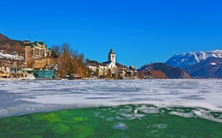 Verschneite Landschaft mit Kirche am zugefrorenen See und Bergen im Hintergrund.