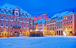 Winterlicher Stadtplatz mit historischen Gebäuden, beleuchtet bei Dämmerung, verschneit.