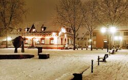 Verschneiter Platz bei Nacht mit beleuchtetem Fachwerkhaus im Hintergrund.