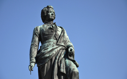 Statue eines historischen Mannes in edler Kleidung aus Bronze vor einem klaren blauen Himmel.