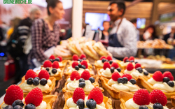 Nahaufnahme von Gebäck mit Himbeeren und Blaubeeren, zwei Personen im Hintergrund.