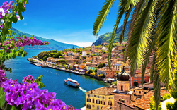 Mediterrane Küstenstadt mit Palmen und blühenden Blumen, im Hintergrund ein See.