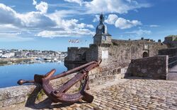 Historische Stadtmauer mit großem Anker, Hafen und Uhrturm im Hintergrund.