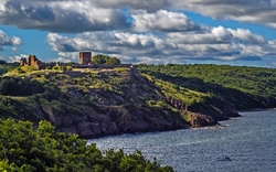 Historische Burg auf einem bewaldeten Hügel mit Blick auf das Meer, bei bewölktem Himmel.