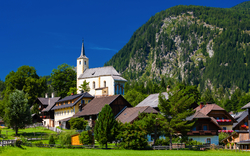 Bergdorf mit Kirche und traditionellen Häusern vor einem bewaldeten Berg.