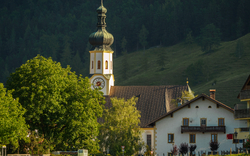 Landschaft mit Kirche, Bäumen und Häusern vor bewaldetem Hügel.