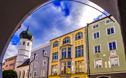 Blick durch einen Torbogen auf bunte Gebäude und einen Kirchturm bei bewölktem Himmel.