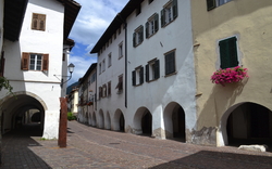 Gasse mit historischen Gebäuden, Arkaden und blühendem Blumenfenster im Sonnenschein.