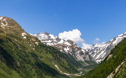 Blick auf ein grünes Tal mit Fluss und schneebedeckten Bergen unter blauem Himmel.