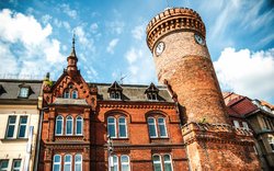 Roter Backsteinturm und historisches Gebäude vor blauem Himmel.