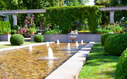 Garten mit Brunnen, umgeben von Hecken und Blumentöpfen, in sommerlicher Umgebung.