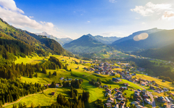 Luftaufnahme eines grünen Tals mit Bergen und einem Dorf bei Sonnenschein.