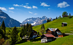 Alpenlandschaft mit Hügeln, Häusern und Bergen im Hintergrund.