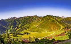 Panorama der Alpen mit grünen Tälern und bewaldeten Berggipfeln unter blauem Himmel.