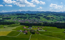 Landschaft mit grünen Feldern, Hügeln, einem Dorf und Bergen im Hintergrund.
