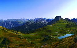 Weite Berglandschaft mit grünen Hügeln, einem See und blauem Himmel.