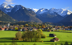 Alpenlandschaft mit Dorf im Tal, grünen Wiesen und schneebedeckten Bergen im Hintergrund.