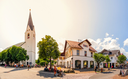 Ortszentrum mit Kirche, Wohnhäusern und Passanten an einem sonnigen Tag.