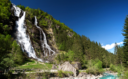 Wasserfall in bergiger Landschaft mit Wald und klarem Himmel.
