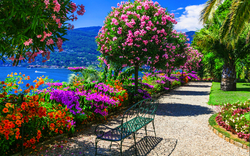 Blumenreicher Gartenweg mit Bank und Blick auf einen See an einem sonnigen Tag.