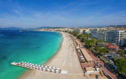 Strand mit Liegen und türkisfarbenem Wasser an der Küste von Cannes, gesäumt von Gebäuden.