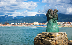 Skulptur von Mutter mit Kind am Meer vor Bergkulisse.